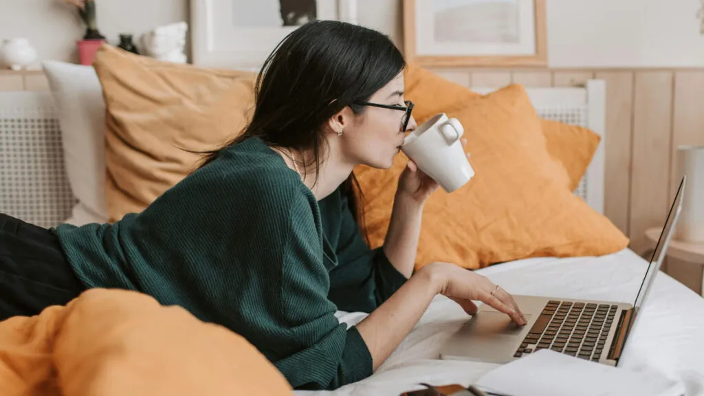Ung kvinna med glasögon som ligger på sängen hemma och dricker kaffe framför en bärbar dator