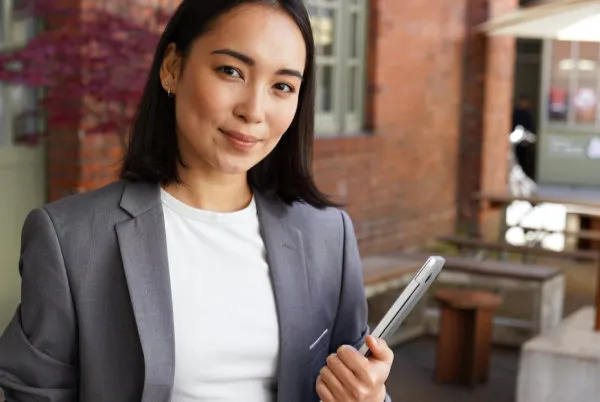 Young woman in a grey blazer holding a tablet outside a brick building