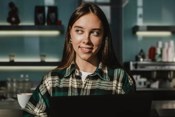 Young woman in a checked shirt smiling at a laptop in a café